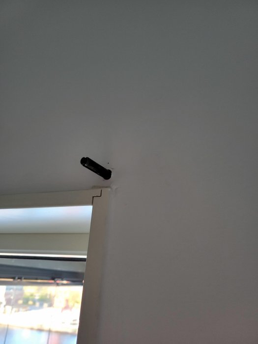 Svart övervakningskamera monterad på vit vägg i ett hörn nära taket, inomhus, med fönster i bakgrunden.