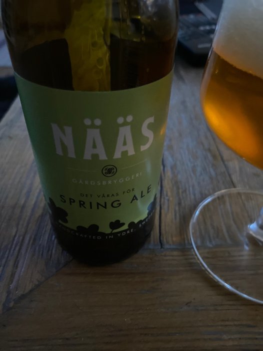 Ölflaska med etikett "NÄÄS Gårdsbryggeri Spring Ale" intill ölglas på ett bord.