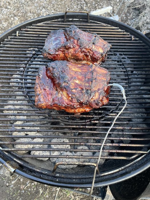 Två stora revbenstycken som grillar på en kolgrill, vackert glaserade, med termometertråd för temperaturkontroll.