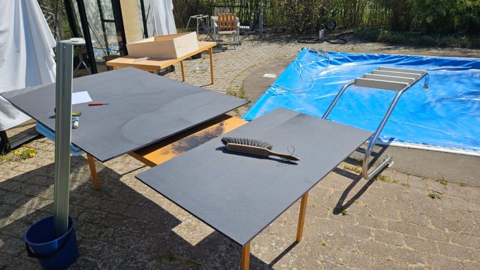 Utomhus med bord, sandpapper, pensel, pool, trappa, soligt väder, arbetsområde för snickeri eller renovering.