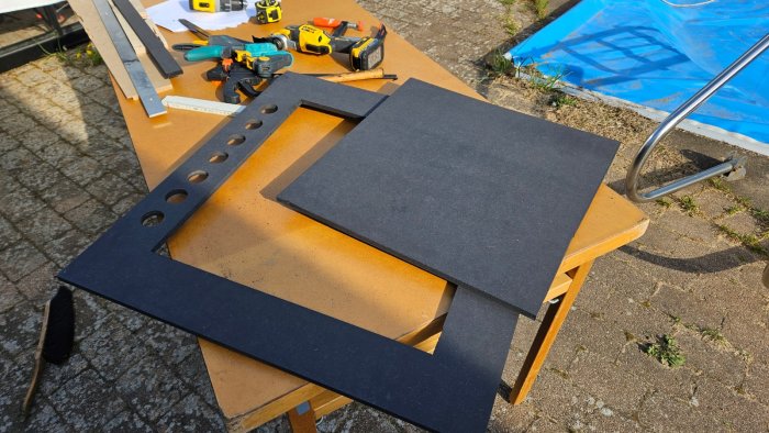 Arbetsbord utomhus med skumplattor, verktyg, och trästycken; DIY-projekt nära pool.