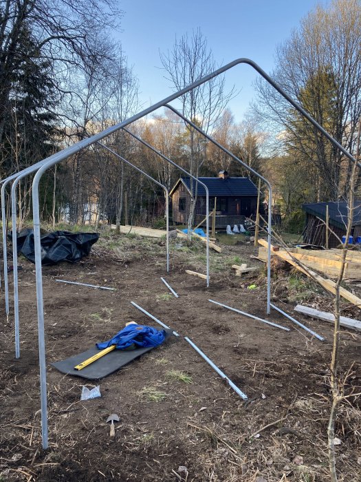 Trädgård; växthus under konstruktion; stålrör; verktyg på marken; träd; hus i bakgrunden.