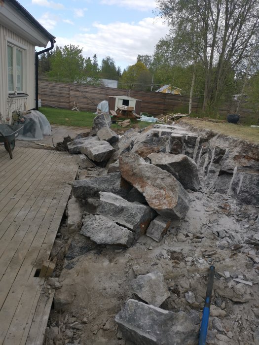 Trädgårdsarbete med stora stenblock, röra, gräsmatta i bakgrunden, skottkärra, träaltan nära hus.