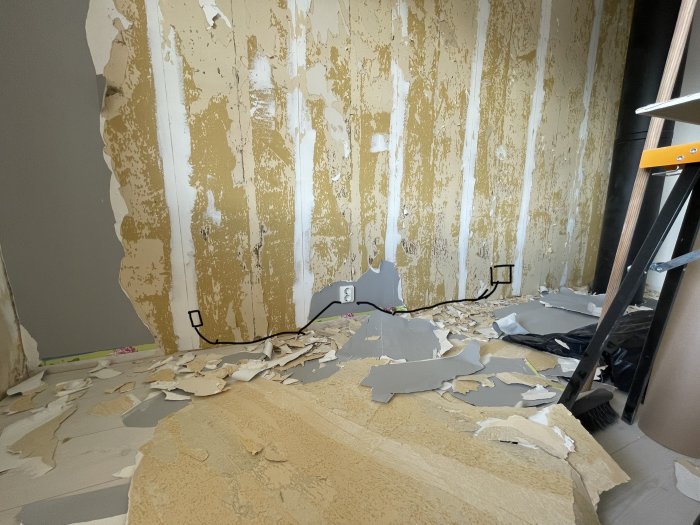 Renoveringsarbete pågår, tapeten tas bort, väggen är skadad, skräp på golvet, stege till vänster.