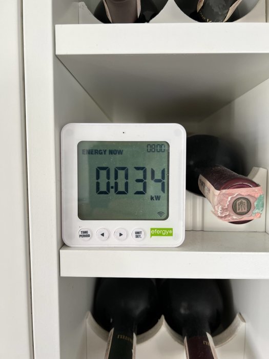 En energimätare visar 0.034 kW förbrukning, placerad på en vit hylla med vinflaskor.