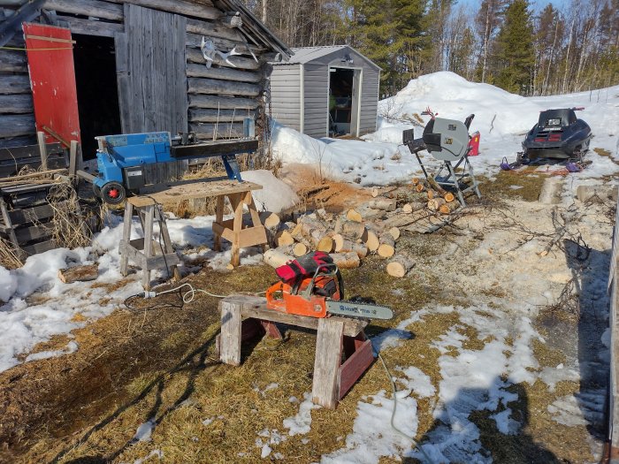 Vedkapningsutrustning utomhus vid lada, motorsåg, vedkap, snöskoter, smältande snö, soligt, träbearbetning.