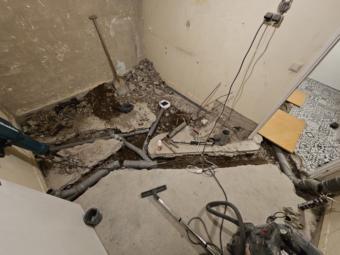 Renovering pågår, uppbrutet golv, synliga rör, verktyg, byggbråte, delvis kaklad vägg.