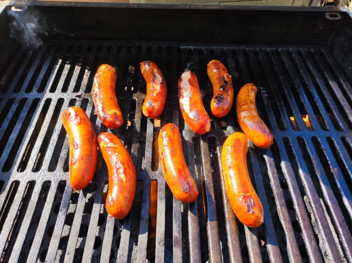 Korvar grillas på en gasolgrill; vissa är karamelliserade och grillyta synlig.