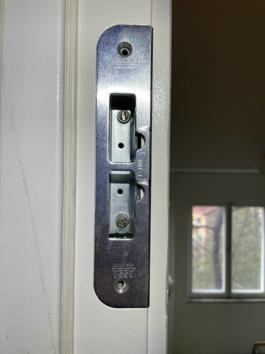 Metallisk dörrlås platta, vit dörrkarm, skruvad, säkerhetsmärke, inomhus, fönster i bakgrunden.