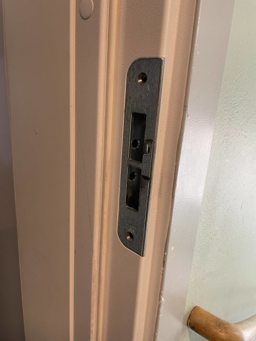 Dörrstolpe med metallisk låsbleck, saknar låscylinder och handtag, vit dörrkarm, del av brun dörrhandtag.