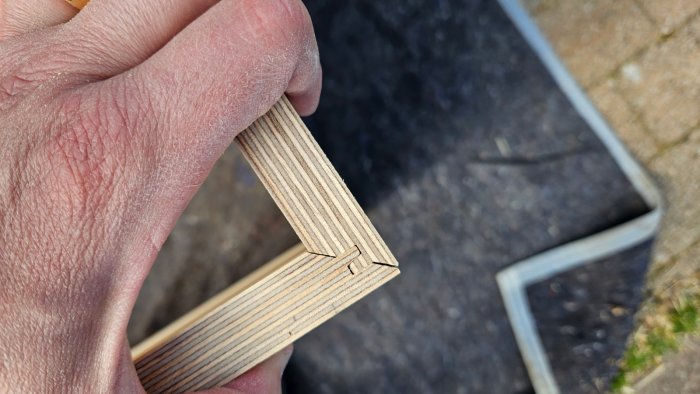 Hand håller plywoodbitar med genomgående fingerskarv mot utomhusbakgrund.