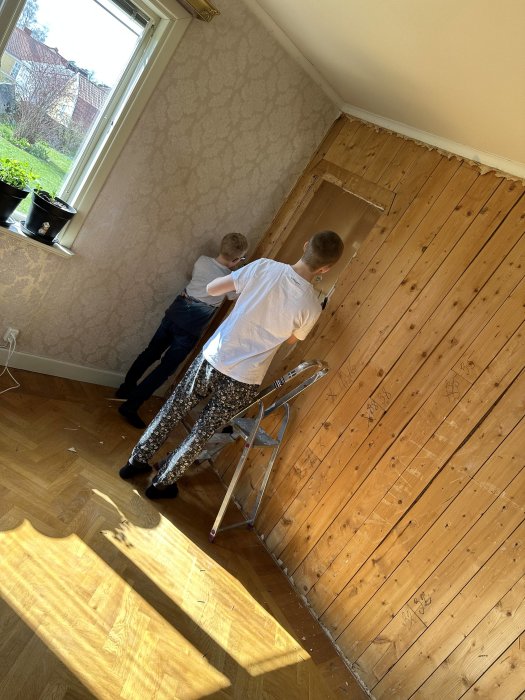 Två personer arbetar med husrenovering i ett rum med träpanel och tapet.