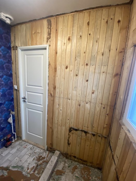 En rumshörna under renovering med träpanelväggar, rörig golv med riven tapet och en vit dörr.