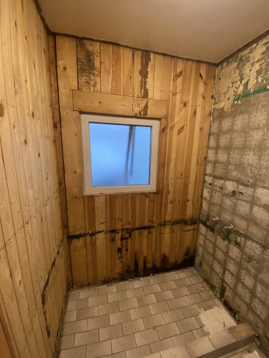 Orenoverat rum med träpaneler och kala väggar, enkelt fönster, fläckigt och ofullständigt, kräver renovering.