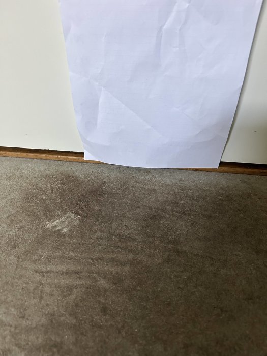 Vitt papper på vägg, smutsigt brunt golv, inomhus, skrynkligt, enkel bakgrund, text längst ner på pappret.