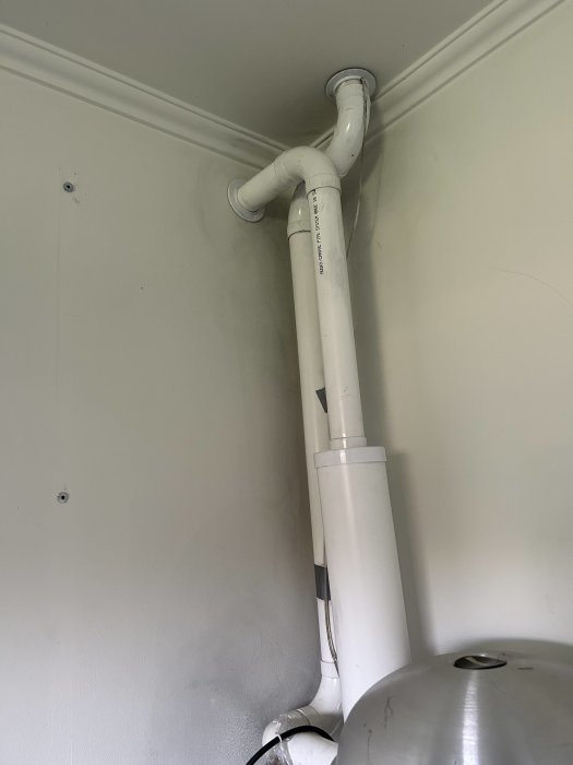 Vit PVC-rörledning löper längs en vägg och tak med synlig böj och anslutning, ovanför metallföremål.