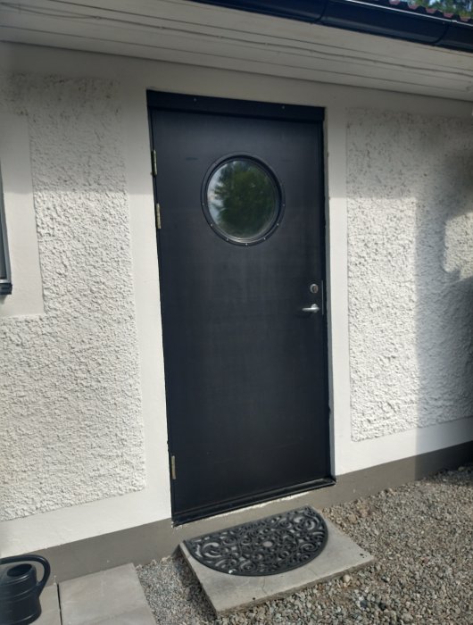 Svart ytterdörr till hus med rund fönstercirkel, dörrmatta och vit putsad vägg.