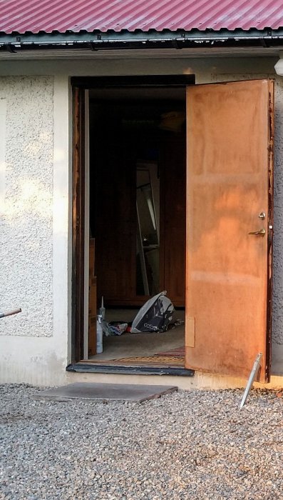 Uppfart med grus, öppen rostig dörr till hus med synlig inredning och rörigt.