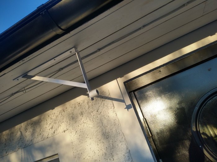 Vit väggfäste för flaggstång, takränna, dörr i solljus, skuggor, klarblå himmel.