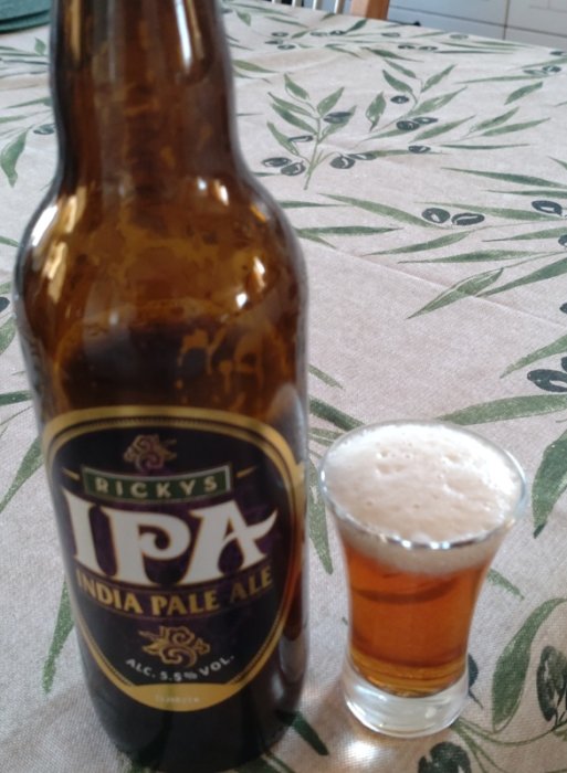 En ölflaska med etikett "IPA" och ett halvfullt glas med skummande öl på ett blommönstrat bord.