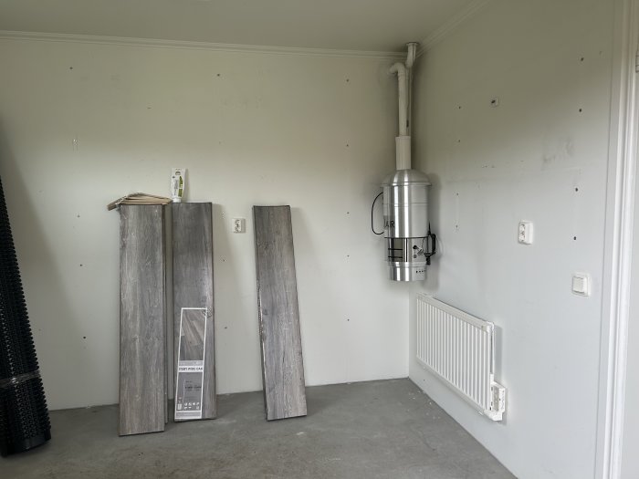 Tomt rum med gråa laminatgolvplankor, värmeelement, vägglampett och en svart matta.