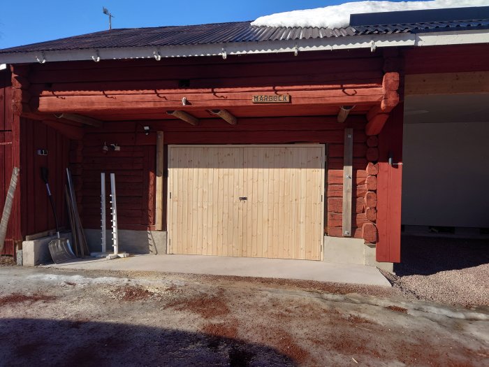 Röd träbyggnad med dubbelgarageport i ljust trä under snötäckt tak. Solig dag.