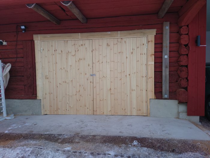 Träräcke för garage på röd stuga, snötäckt mark, betonggrund, dagtid.