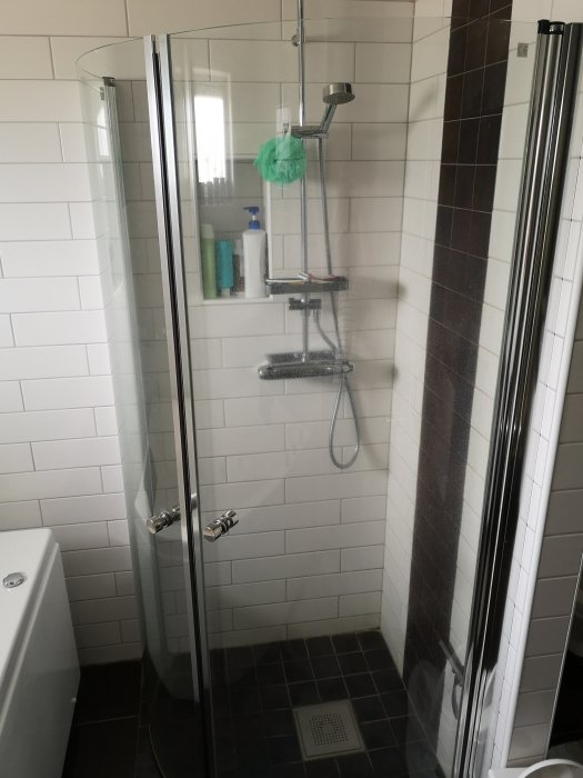 Modernt badrum med duschhörna, genomskinliga glasdörrar, vita och mörkbruna kakelväggar, hygienprodukter och duschmunstycke.