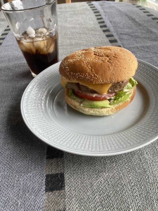 Hamburgare med sallad, tomat, ost och kött på tallrik bredvid iskall cola.