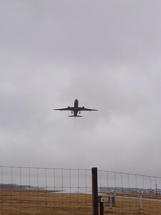 Flygplan landar, grå himmel, flygplatsstaket, fält i förgrunden, dimmig dag.