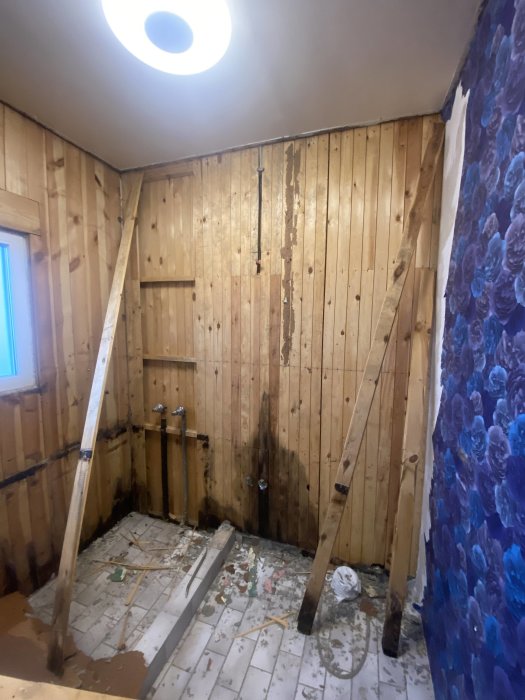Renoveringsarbete pågår, avskalat rum, träväggar, avbladad tapet, blottade rör, skräp på golvet, taklampa.