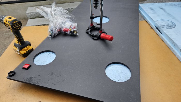 Arbetsyta med borr, knappsats, hål, plastpåse, skruvklammer och borrställ på gata.