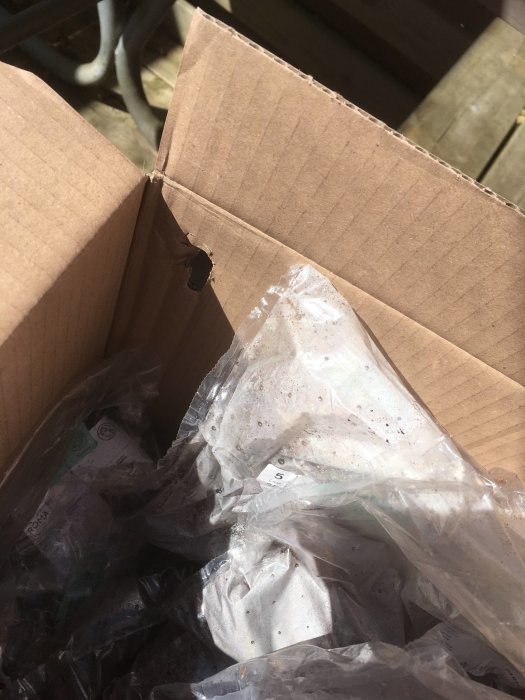 Well-lit cardboard box with torn corner, containing plastic-wrapped items, possibly soil or sand.