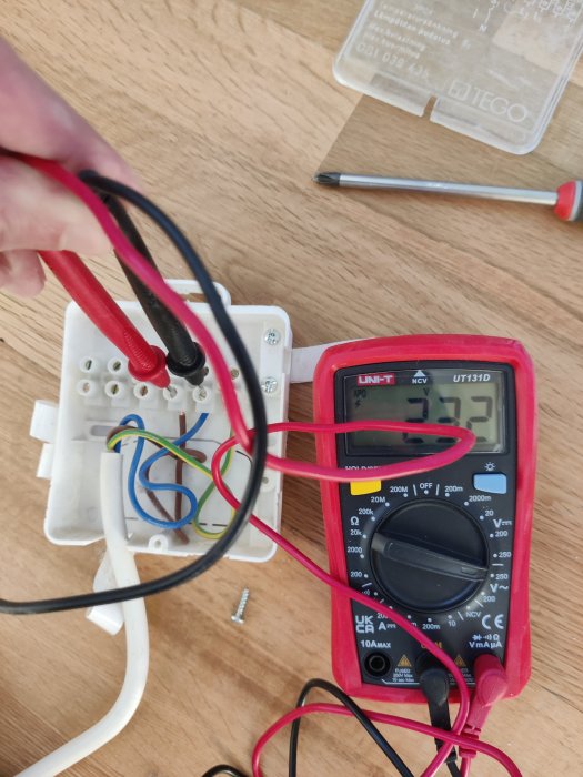 Mätning av elektrisk spänning i vägguttag med en digital multimeter. Kablar och skruvmejsel synliga.