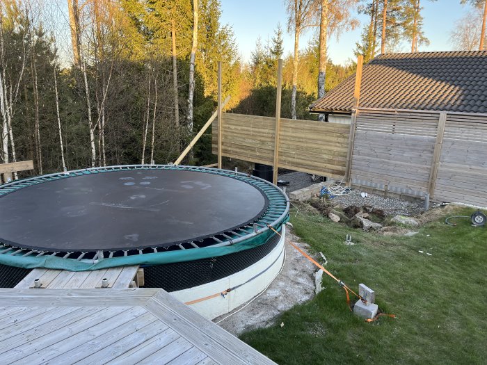 Trädgård med studsmatta, träterrass, staketbygge, träd och gräsmatta i soligt väder.