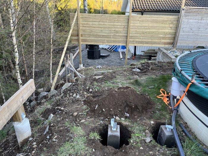 Utomhusarbetsplats med grävning, stolpar, trädäck, pool, och byggmaterial i skymning.
