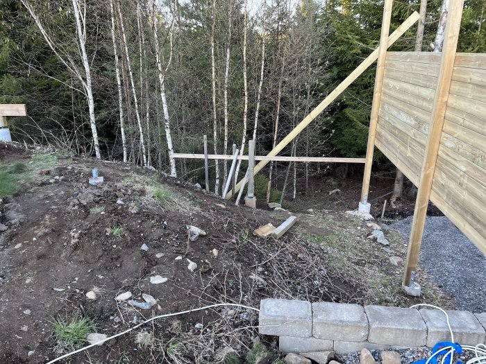 Trädgårdsarbete under konstruktion: lerjord, stenmur, trästöttor, skog i bakgrunden, delvis byggd staket.