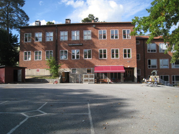 Tvåvånings rödtegelbyggnad, skylt "LJUMSVIKSSKOLAN", soligt, gröna träd, skuggor, utebord, cykelställ, skolområde.