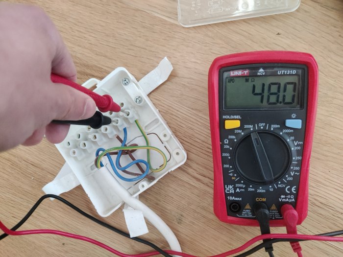 Hand använder multimeter för att mäta spänning på elinstallation, skruvmejsel, ledningar och skyddsplast synlig.