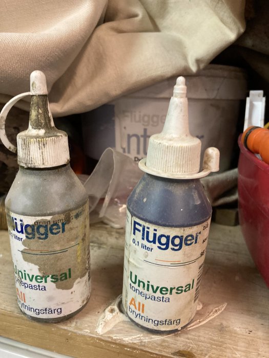 Två använda flaskor med toningspasta märkta "Flügger" på ett dammigt hyllplan.