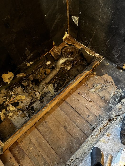 Nedrivet golv visar gamla rörledningar och skräp i ett förfallet utrymme.