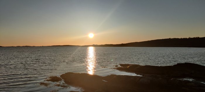 Solnedgång över havet, reflektion på vatten, klippor i förgrunden, lugnt och stillsamt landskap.