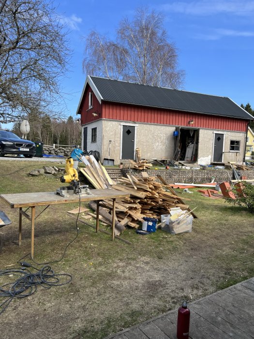 Renoveringsprojekt med material och verktyg framför röd och grå byggnad, soligt, bostadshus i bakgrunden.