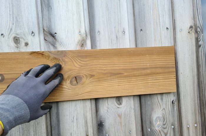 Hand i arbets handske håller ett ommålat träbräde mot en målad trävägg.
