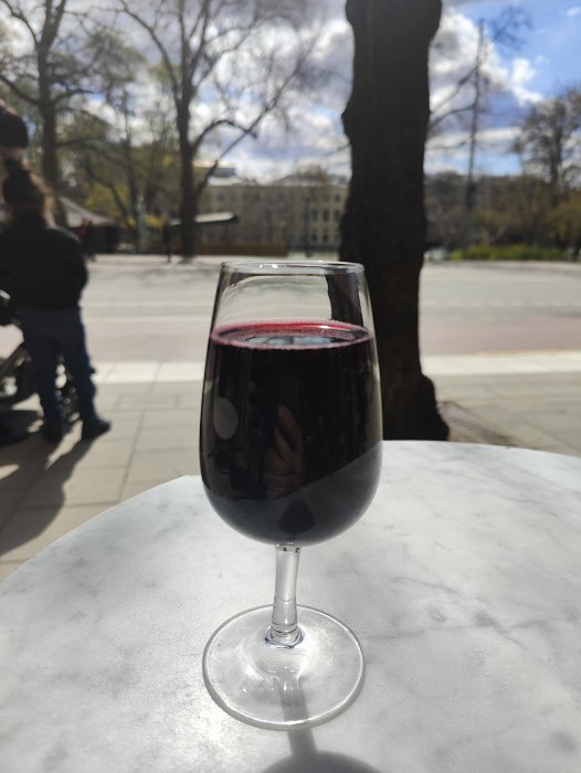Ett glas vin på ett bord utomhus, en suddig bakgrund med träd och personer i solljus.
