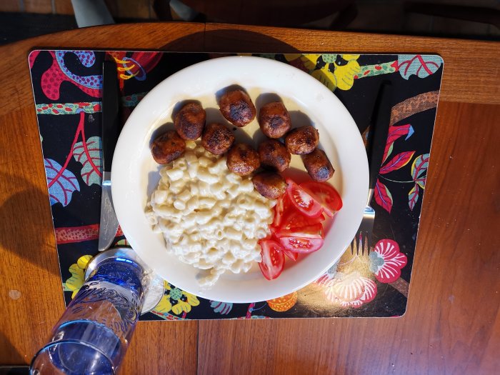 Makaroner, köttbullar, tomatskivor på vit tallrik, vattenglas, färgglad bordstablett, bestick, träbord.