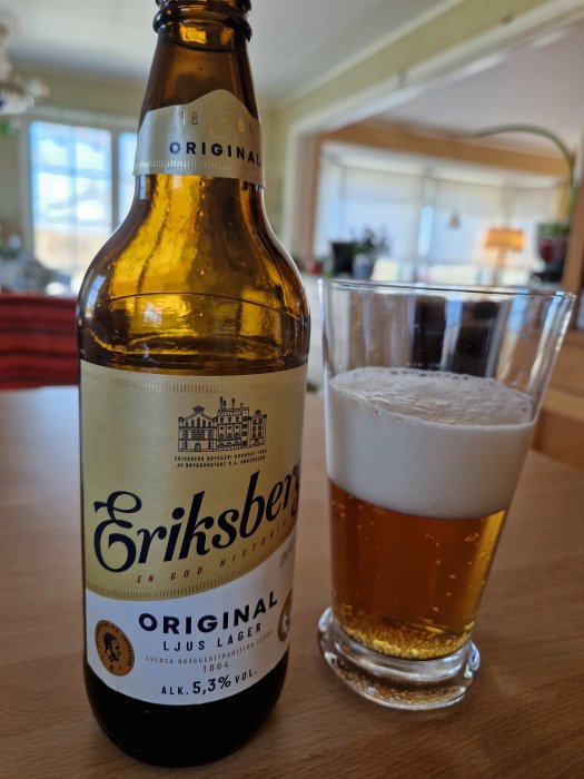 En ölflaska märkt 'Eriksberg' bredvid ett glas med öl, halvt fullt, skumtäcke, inomhusmiljö.