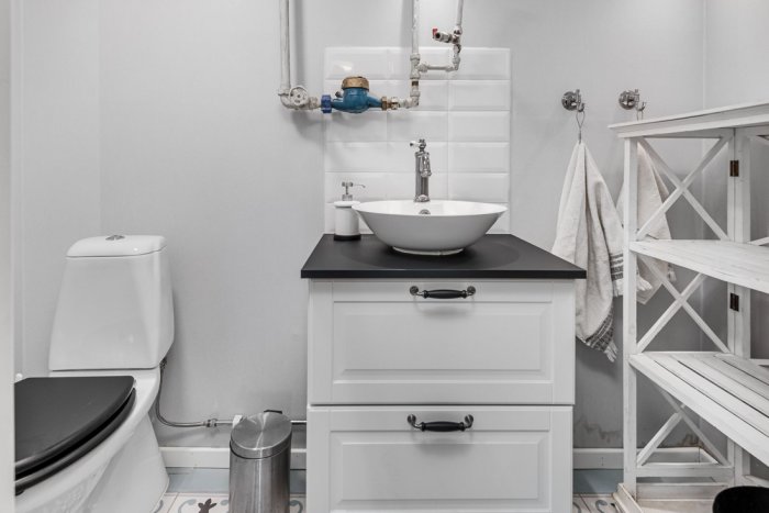Modernt badrum med vit toalett, handfat på förvaringsenhet, handduksställ, och vitkaklade väggar.