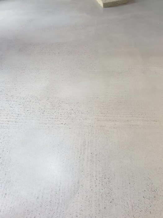 Ett ljust, grått golv med små fläckar, möjligen betong. Sparsmakad inredning, avskalat utrymme.