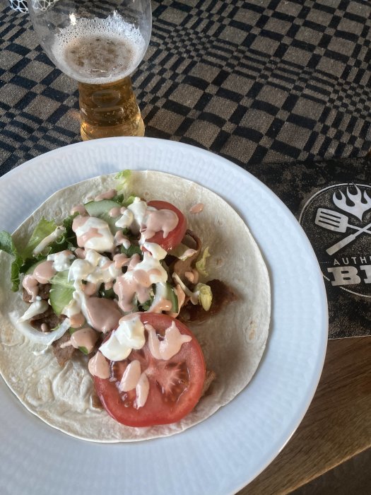 Öppen tortilla med sallad, tomat, sås; halvtomt ölglas; rutig duk.